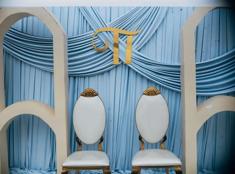 Couple sweetheart reception stage with powder blue pleated fabric backdrop, gold monogram emblem, modern cream arch frames, and gold-trimmed white oval chairs