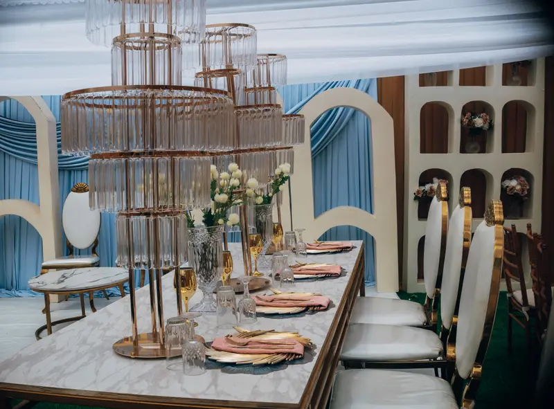 Opulent wedding banquet tablescape featuring a tiered crystal candelabra chandelier centerpiece, gold beaded charger plates, blush napkins, and powder blue drapery